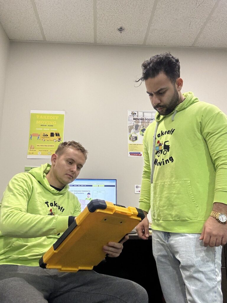 Two men in green hoodies review a yellow tablet in an office with moving company posters. Takeoff-Moving Toronto